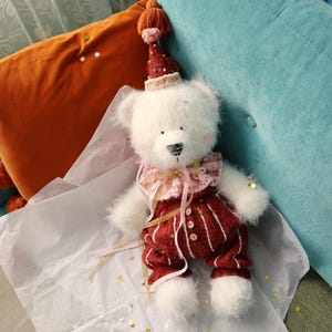 Puede incluir: Un oso de peluche blanco con un atuendo a rayas rojas y blancas y un gorro de fiesta. El oso tiene un cuello rosa con volantes y está sobre papel blanco con confeti de estrellas doradas. El fondo incluye cojines naranjas y turquesas.