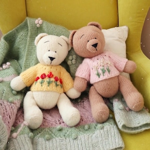 May include: Two handmade teddy bears. One is cream-colored with a yellow sweater embroidered with red flowers. The other is brown with a pink sweater featuring floral embroidery. They rest on a green and pink knitted blanket.