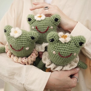May include: Three handmade crochet frog dolls. Each frog is green with black eyes, a red stitched smile, and a white flower on its head. The frogs wear ruffled collars in shades of pink and cream.