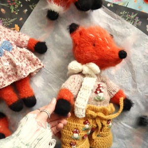 Pode incluir: Brinquedos de pelúcia de raposa laranja feitos à mão com patas e orelhas pretas. Uma raposa usa um suéter rosa, shorts amarelos com bordados de cogumelos e um cachecol branco. Outras raposas são visíveis no fundo.