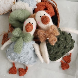 May include: Two handmade knitted goose dolls with white bodies, orange beaks and feet, and black eyes. One goose wears a light blue dress with bobbles and a green bow. The other wears a green textured dress and a brown bow. A basket of yarn is in the background.