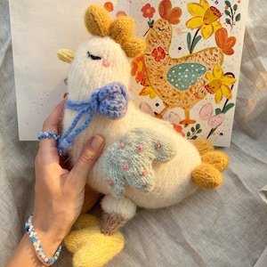 May include: A handmade, cream-coloured knitted chicken toy with yellow detailing, a blue bow, and pale blue wings with pink spots. A greeting card with a chicken illustration and Russian text is in the background.