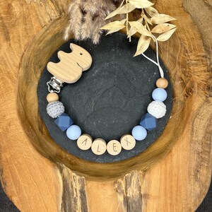Könnte beinhalten: Ein blauer und grauer Silikon- und Holz-Beißring mit dem Namen "ALEX" auf den Holzperlen. Der Beißring hat einen Holzclip in Tierform.