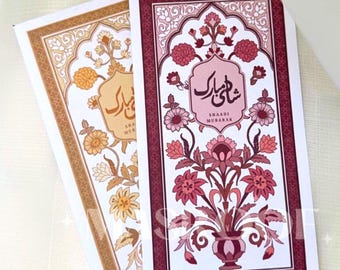 May include: Two rectangular cards with floral designs. One card has a gold border and a floral pattern in gold and cream. The other card has a burgundy border and a floral design in pink and burgundy. The text "SHAADI MUBARAK" is visible.