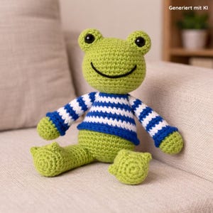 May include: A handmade green crochet frog doll with a smiling face, black eyes, and a blue and white striped sweater. The frog is sitting on a beige sofa. The text "Generiert mit KI" is in the top right corner.