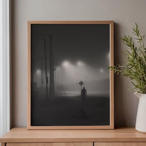 Könnte beinhalten: Ein gerahmter Wandkunstdruck mit einer monochromen, nebligen Nachtszene, in der eine einsame Gestalt auf einer dunklen Straße geht. Die Komposition umfasst Strommasten, leuchtende Straßenlaternen und ein rautenförmiges Verkehrsschild in Grautönen.
