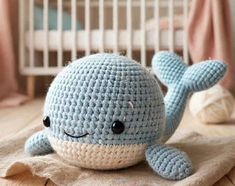 May include: A light blue and cream-colored crocheted whale plush toy with a rounded body, a wide tail, two small side fins, and two black plastic eyes. The whale features a textured yarn surface and a stitched smile on its front face.