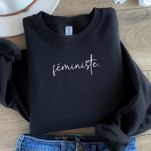 Könnte beinhalten: Schwarzer Pullover mit dem Wort "féministe" in weißer Kursivschrift.