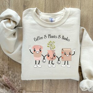 Könnte beinhalten: Ein beiger Pullover mit dem Aufdruck "Coffee & Plants & Books" auf der Vorderseite. Der Text ist von Cartoon-Illustrationen einer Kaffeetasse, einer Topfpflanze und einem Buch umgeben, die jeweils ein lächelndes Gesicht haben.