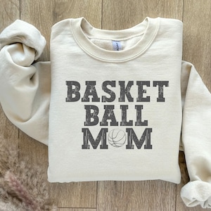 May include: Cream-colored sweatshirt with the text "BASKET BALL MOM" in dark gray, with a basketball graphic. The sweatshirt is folded, with the sleeves and collar visible. The fabric appears soft and comfortable.