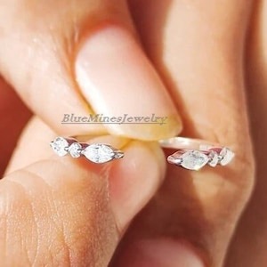 Può includere: Un anello d'argento con tre piccole pietre chiare a forma di marquise incastonate in una fascia delicata. L'anello è aperto e regolabile.