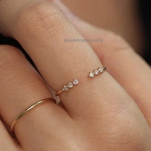 Puede incluir: Un delicado anillo de oro con una fila de pequeños diamantes engastados en forma de curva. El anillo está abierto por un lado y se lleva en un dedo.