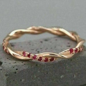 Puede incluir: Un delicado anillo de banda de oro con un diseño trenzado y una fila de pequeñas piedras preciosas rojas.