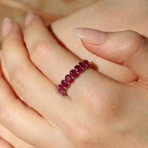May include: A silver ring featuring a series of oval-cut, deep red gemstones. The gemstones are set closely together, creating a continuous band of color. The ring is shown on a finger, highlighting the vibrant red of the stones.
