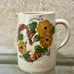 May include: A white ceramic pitcher with a hand-painted design of brown and yellow flowers with green leaves. The pitcher has a brown handle and a signature on the bottom.