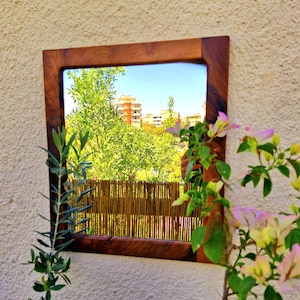 Könnte beinhalten: Ein quadratischer Spiegel mit Holzrahmen, der eine Szene im Freien widerspiegelt. Der Rahmen ist warmbraun. Die Reflexion zeigt grüne Bäume, einen Bambuszaun und einen blauen Himmel. Rosa und weiße Blumen und grüne Blätter umrahmen den Spiegel.