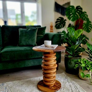 Può includere: Un tavolino rotondo in legno con una base a piedistallo impilata a forma di anelli in legno chiaro. Il piano del tavolo è circolare con una finitura liscia in legno scuro. Una piccola tazza in ceramica bianca e beige poggia su una rivista.