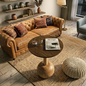 Op de afbeelding: Een ronde houten bijzettafel met een bolvormige voet, voorzien van een glazen mok en een boek met de titel design op het oppervlak. De tafel staat voor een gecapitonneerde bruine leren bank met kussens met patronen en een crèmekleurige poef.
