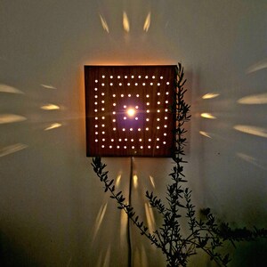 May include: A wooden wall lamp with a square shape and a grid of holes. The light shines through the holes, creating a pattern of light on the wall. The lamp is mounted on a white wall.