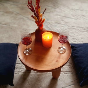 May include: Two glasses of red wine, a lit candle, and a vase with dried flowers on a wooden table with two blue cushions on the floor.