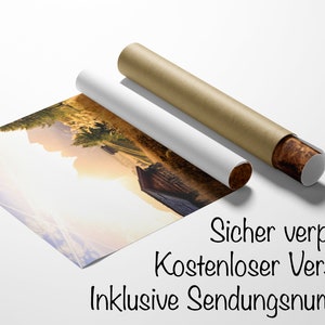 May include: A rolled up print of a landscape photograph featuring a wooden cabin in a field with mountains in the background. The text "Sicher verpackt Kostenloser Versand Inklusive Sendungsnummer" is printed on the bottom of the image.