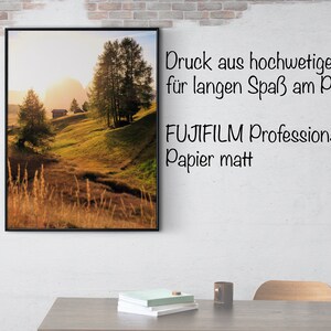 May include: A framed landscape photograph of a grassy hillside with a small wooden cabin in the distance. The sun is setting behind the hills, casting a warm glow over the scene. The text "Druck aus hochwertigem Papier f&uuml;r langen Spa&szlig; am Poster FUJIFILM Professional Papier matt" is printed below the image.
