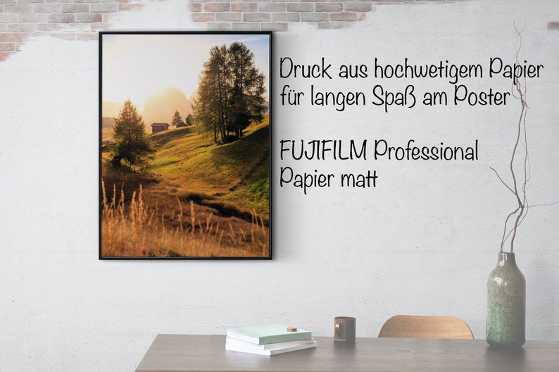 May include: A framed landscape photograph of a grassy hillside with a small wooden cabin in the distance. The sun is setting behind the hills, casting a warm glow over the scene. The text "Druck aus hochwertigem Papier f&uuml;r langen Spa&szlig; am Poster FUJIFILM Professional Papier matt" is printed below the image.