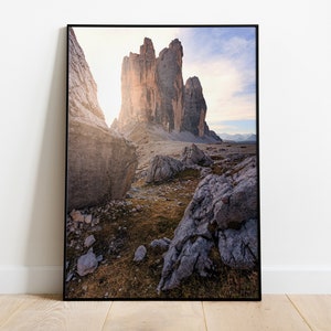 May include: A framed photograph of a mountain range with a dramatic rock formation in the center. The sun is shining through the rocks, creating a warm glow. The foreground is a rocky landscape with some grass and small rocks.