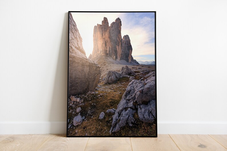 May include: A framed photograph of a mountain range with a dramatic rock formation in the center. The sun is shining through the rocks, creating a warm glow. The foreground is a rocky landscape with some grass and small rocks.