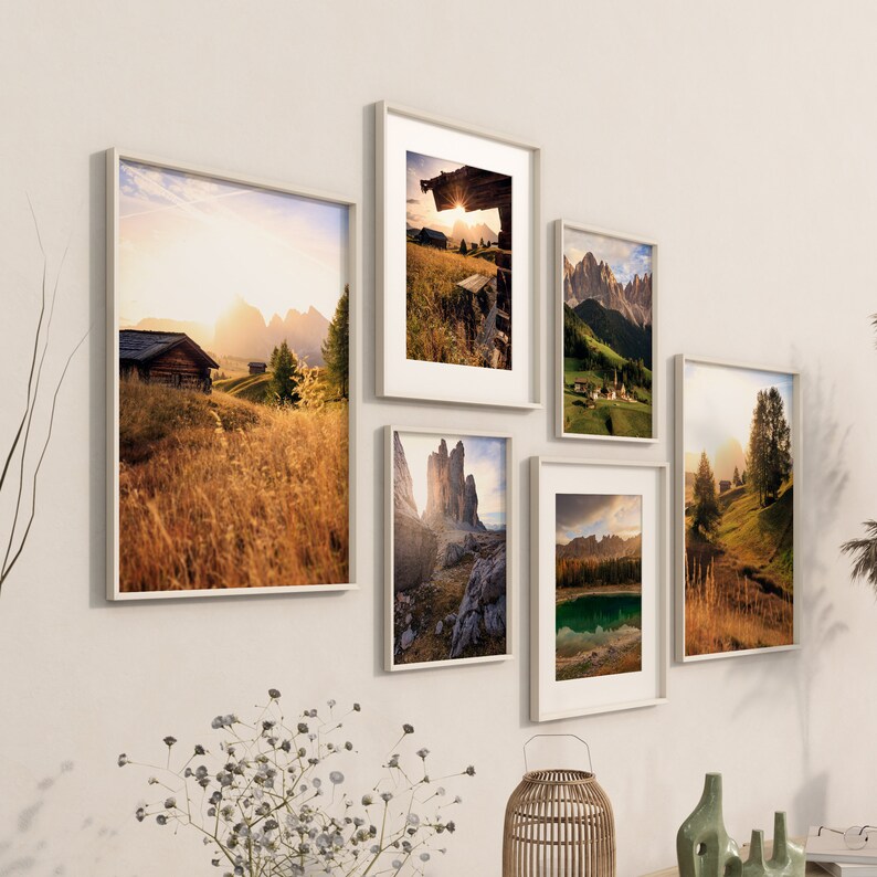 May include: A gallery wall display of six framed landscape photographs featuring mountain ranges, lakes, and forests. The photos are in white frames and are arranged in a staggered pattern on a white wall.