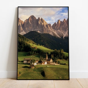 May include: A framed photograph of a mountain range with a small village nestled in the valley below. The village has a church with a tall steeple and several houses. The mountains are a light brown color and the valley is green.
