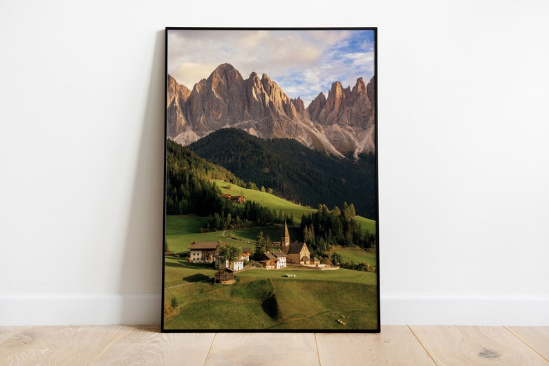 May include: A framed photograph of a mountain range with a small village nestled in the valley below. The village has a church with a tall steeple and several houses. The mountains are a light brown color and the valley is green.