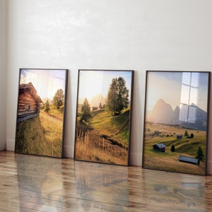 May include: Three framed landscape photographs of a mountainous region with rolling hills, green grass, and small wooden houses. The photos are hung on a white wall.