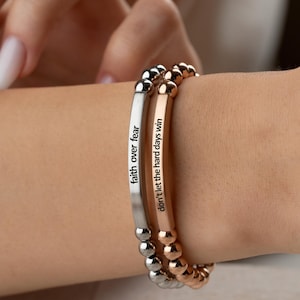 May include: Two beaded bracelets are worn on a wrist. One is silver-toned with the words "faith over fear" engraved on it. The other is rose gold-toned with the words "don't let the hard days win" engraved on it. The bracelets are a stylish accessory.