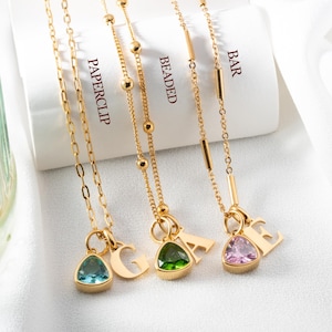 May include: Three gold necklaces with gemstone pendants and initial charms. The necklaces are labeled "PAPERCLIP", "BEADED", and "BAR". Each necklace features a different colored gemstone: blue, green, and pink. A bottle of iced tea is in the background.