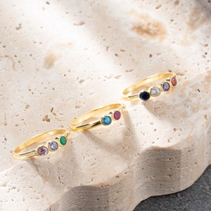 May include: Four gold rings with different colored gemstones. The rings are arranged in a row on a light brown textured surface.
