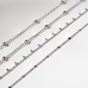 May include: Four delicate silver necklaces with varying designs. One features small, evenly spaced beads, while others have a mix of larger and smaller beads. The chains are displayed against a white background, highlighting their shine.