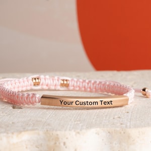 May include: A pink braided bracelet with a gold bar that says "Your Custom Text".