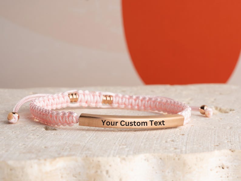 May include: A pink braided bracelet with a gold bar that says "Your Custom Text".