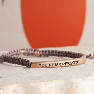 May include: A gray braided bracelet with a gold bar charm that reads "You're My Person".