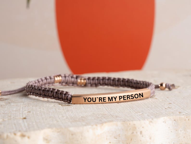 May include: A gray braided bracelet with a gold bar charm that reads "You're My Person".