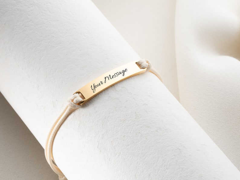 May include: A gold-colored bracelet with the text "Your Message" engraved on a rectangular plate. The plate is attached to the bracelet with cream-colored cords. The bracelet is displayed on a white, textured surface.