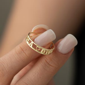 May include: A gold ring with the names "OLA" and "DYO" spelled out around the band, separated by a heart symbol.