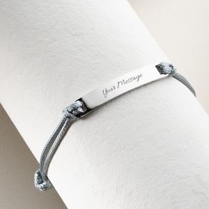 May include: A gray bracelet with a rectangular silver plate engraved with the words "Your Message" in cursive. The bracelet is made of gray rope and is knotted on each side of the plate.