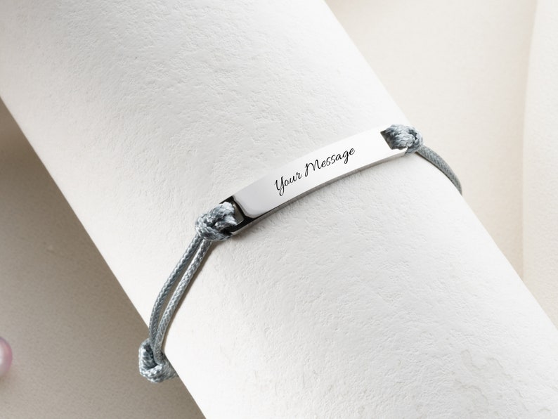 May include: A gray bracelet with a rectangular silver plate engraved with the words "Your Message" in cursive. The bracelet is made of gray rope and is knotted on each side of the plate.