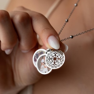 May include: Silver locket necklace with a decorative openwork pendant and three circular photo pendants. The locket pendant features a geometric design with small, clear gemstones. The necklace chain has small, round beads.