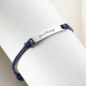 May include: A silver bracelet with a navy blue cord. The bracelet features a rectangular plate with the words "Your Message" engraved on it. The cord is knotted at both ends of the plate.