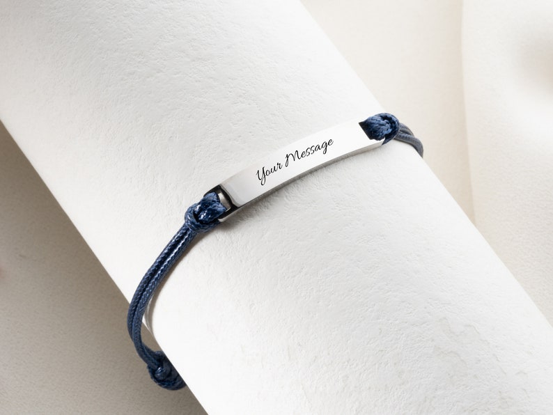 May include: A silver bracelet with a navy blue cord. The bracelet features a rectangular plate with the words "Your Message" engraved on it. The cord is knotted at both ends of the plate.