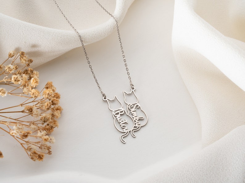 Op de afbeelding: Zilveren ketting met twee katten-silhouetten, elk met een naam erin gegraveerd: "Buster" en "Simba".