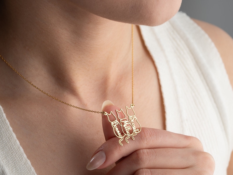 Op de afbeelding: Gouden ketting met een ketting en vier kattenhangers. Elke hanger heeft een gegraveerde naam: Scout, Millie en twee namen die niet zichtbaar zijn op de afbeelding.